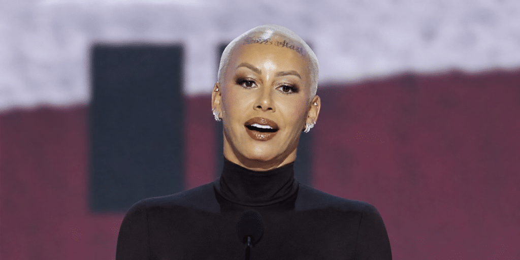 Woman with a platinum blonde buzz cut speaking at a podium on stage, wearing a black turtleneck.