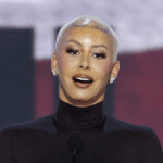Woman with a platinum blonde buzz cut speaking at a podium on stage, wearing a black turtleneck.