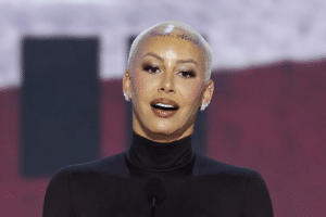 Woman with a platinum blonde buzz cut speaking at a podium on stage, wearing a black turtleneck.