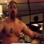 Shirtless man with arms outstretched addressing a group in a dim room, Pod Save America logo visible in the corner.
