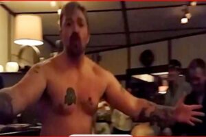 Shirtless man with arms outstretched addressing a group in a dim room, Pod Save America logo visible in the corner.