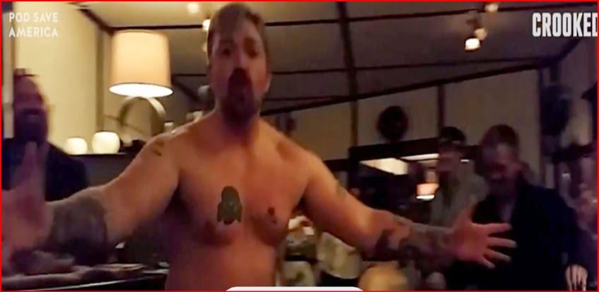 Shirtless man with arms outstretched addressing a group in a dim room, Pod Save America logo visible in the corner.