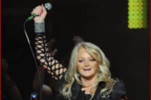 Blonde female singer on stage raising a microphone above her head, wearing a black outfit with lattice sleeves.