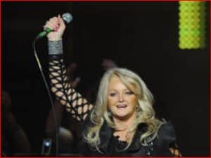 Blonde female singer on stage raising a microphone above her head, wearing a black outfit with lattice sleeves.