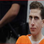 Man in an orange prison jumpsuit seated indoors, looking toward the camera; a blurred figure stands behind him.