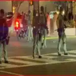 Line of armed police standing across a crosswalk at night, blocking a street; protesters and bystanders in background.