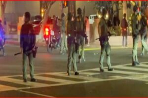 Line of armed police standing across a crosswalk at night, blocking a street; protesters and bystanders in background.