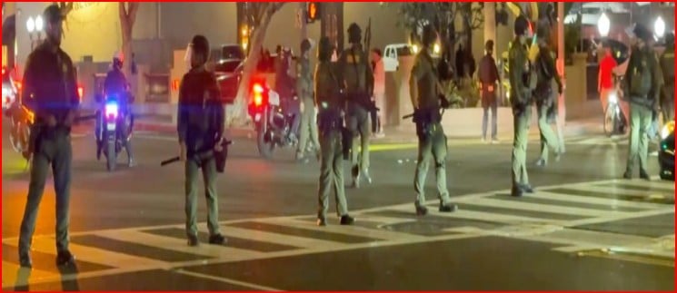 Line of armed police standing across a crosswalk at night, blocking a street; protesters and bystanders in background.