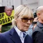 Woman in a navy blazer and sunglasses walks past protesters holding bright yellow signs that read 'Pay Up Deadbeat'.