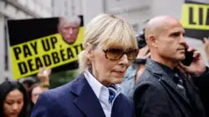 Woman in a navy blazer and sunglasses walks past protesters holding bright yellow signs that read 'Pay Up Deadbeat'.
