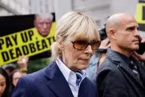 Woman in a navy blazer and sunglasses walks past protesters holding bright yellow signs that read 'Pay Up Deadbeat'.