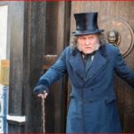 Street performer in a long blue coat and top hat uses a cane as he steps from a dark doorway, while photographers and onlookers watch.