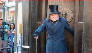 Street performer in a long blue coat and top hat uses a cane as he steps from a dark doorway, while photographers and onlookers watch.