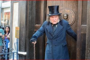 Street performer in a long blue coat and top hat uses a cane as he steps from a dark doorway, while photographers and onlookers watch.