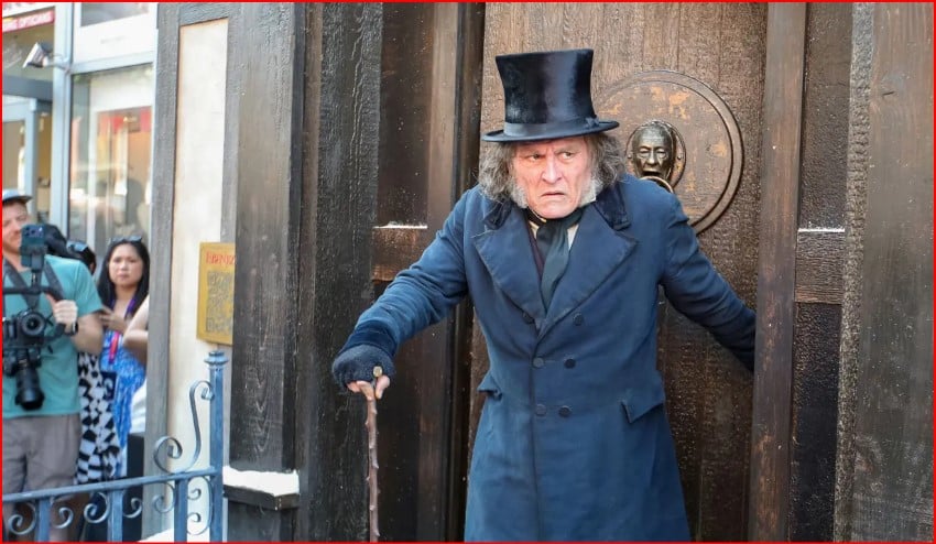 Street performer in a long blue coat and top hat uses a cane as he steps from a dark doorway, while photographers and onlookers watch.