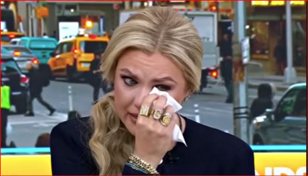 Blonde woman wiping tears with a tissue during a TV interview on a busy city street.