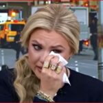 Blonde woman wiping tears with a tissue during a TV interview on a busy city street.