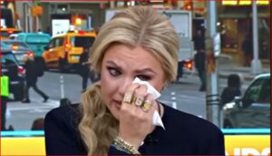 Blonde woman wiping tears with a tissue during a TV interview on a busy city street.