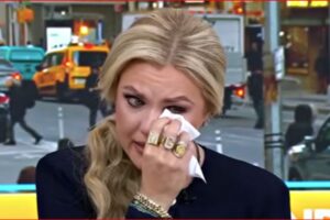 Blonde woman wiping tears with a tissue during a TV interview on a busy city street.