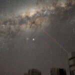 Star-filled night sky with the Milky Way arc and a red laser beam emitting from a rooftop observatory toward the stars.