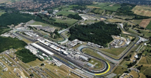 Aerial view of a winding race track with pit buildings and spectator areas amid green fields and tents nearby at a motorsport complex.