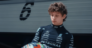 Young race car driver in a dark Petronas racing suit, looking down while holding a helmet near a garage wall.