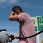 Man in a pink shirt pumps fuel into a dark SUV at a gas station under a blue sky, adjusting his hair as he refuels.