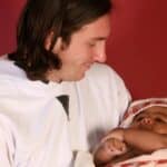 Father cradling a newborn wrapped in a blanket against a red background, both looking down at the baby with a gentle smile