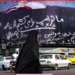 Busy city street with a large curved billboard covered in white cursive slogans, including ‘TRUMP’, as a woman in a black chador walks by among passing cars and motorcycles.