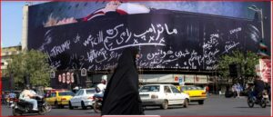 Busy city street with a large curved billboard covered in white cursive slogans, including ‘TRUMP’, as a woman in a black chador walks by among passing cars and motorcycles.