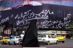 Busy city street with a large curved billboard covered in white cursive slogans, including ‘TRUMP’, as a woman in a black chador walks by among passing cars and motorcycles.