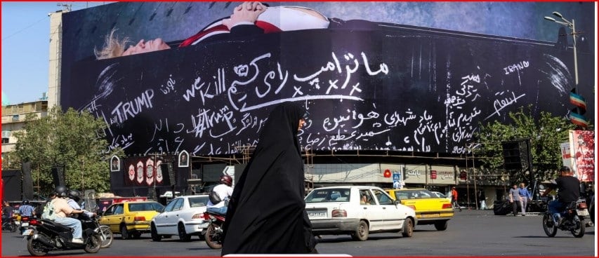 Busy city street with a large curved billboard covered in white cursive slogans, including ‘TRUMP’, as a woman in a black chador walks by among passing cars and motorcycles.