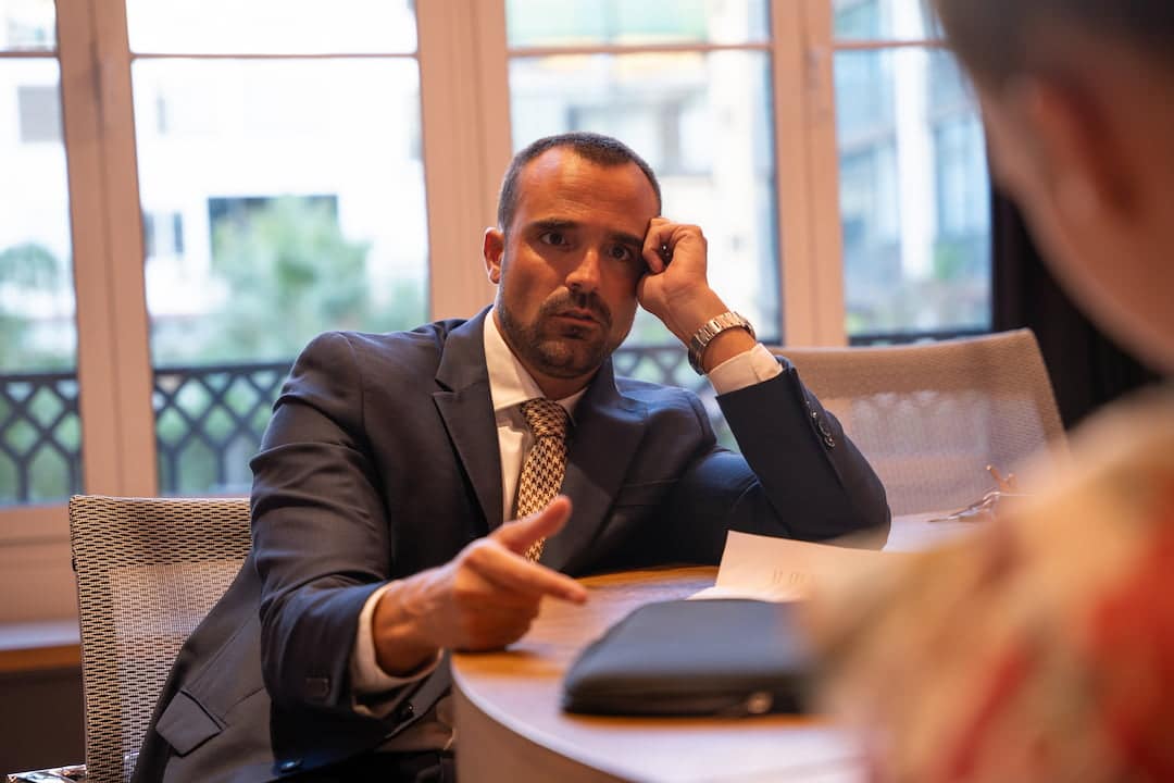 Man in a dark suit sits at a round table in a bright office, pointing his finger while talking to someone off-camera.
