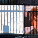 Banner on a fence showing a smiling woman on the right and white panels filled with handwritten messages and a red heart on the left—memorial-style display.