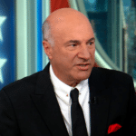 Man in a black suit and white shirt speaking on a television set with blue paneling and star motifs in the background.