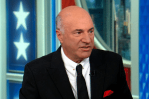 Man in a black suit and white shirt speaking on a television set with blue paneling and star motifs in the background.