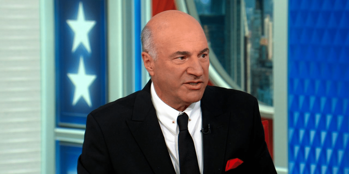 Man in a black suit and white shirt speaking on a television set with blue paneling and star motifs in the background.