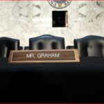 Wooden nameplate reading 'MR. GRAHAM' on a black-draped table in a courtroom, with black chairs and microphones in the background.