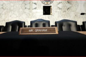 Wooden nameplate reading 'MR. GRAHAM' on a black-draped table in a courtroom, with black chairs and microphones in the background.