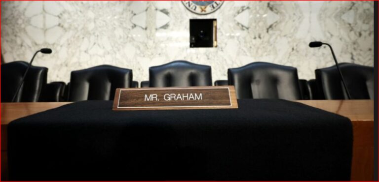 Wooden nameplate reading 'MR. GRAHAM' on a black-draped table in a courtroom, with black chairs and microphones in the background.