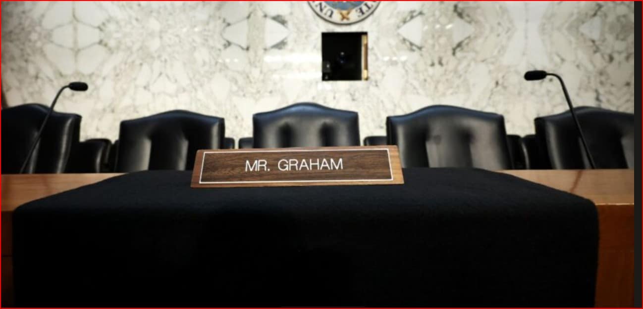Wooden nameplate reading 'MR. GRAHAM' on a black-draped table in a courtroom, with black chairs and microphones in the background.