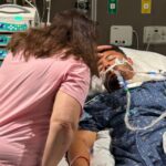 Lonn Hartman lies in an intensive care hospital bed surrounded by medical equipment while a loved one comforts him.
