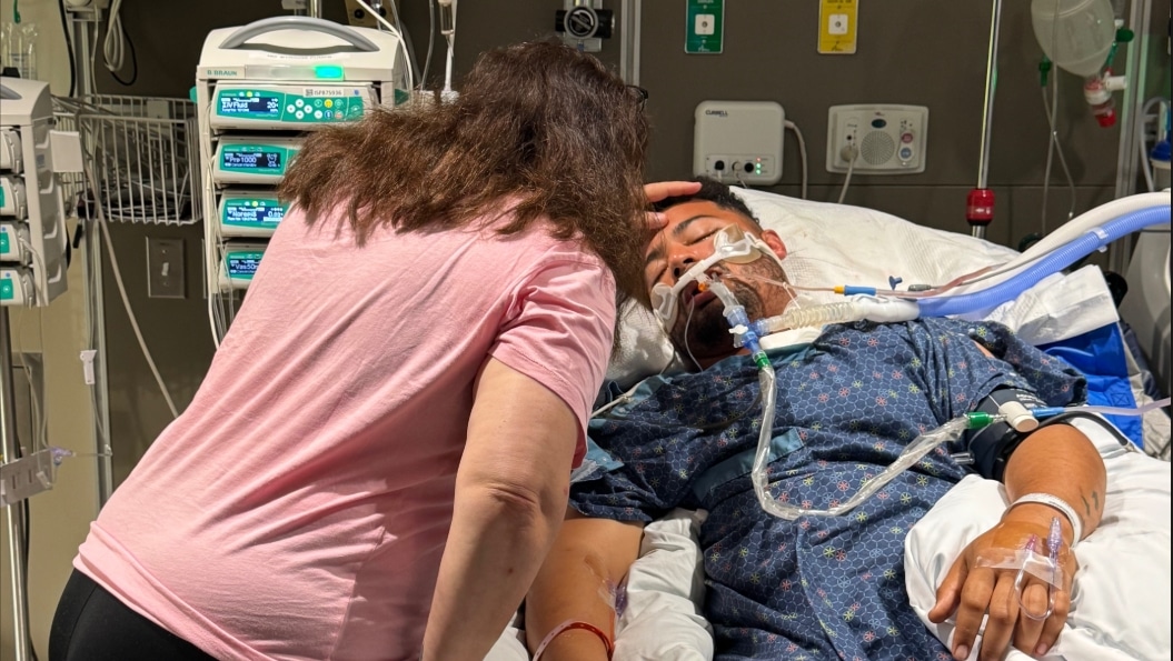 Lonn Hartman lies in an intensive care hospital bed surrounded by medical equipment while a loved one comforts him.