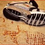Running shoe on a dusty beige surface with red footprints nearby, suggesting bloodstains.