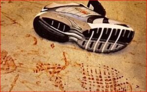 Running shoe on a dusty beige surface with red footprints nearby, suggesting bloodstains.