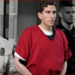 Man in a red jail jumpsuit escorted by two uniformed guards in a hallway