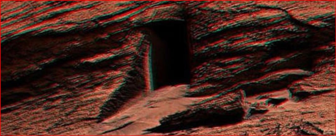 Rocky desert terrain with a dark cave opening carved into the red rock wall (anaglyph 3D effect).