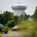 Water tower with Plymouth branding rises above a suburban area, seen behind trees and parked cars on a road in the foreground.
