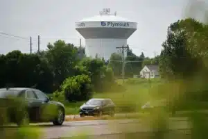 Water tower with Plymouth branding rises above a suburban area, seen behind trees and parked cars on a road in the foreground.