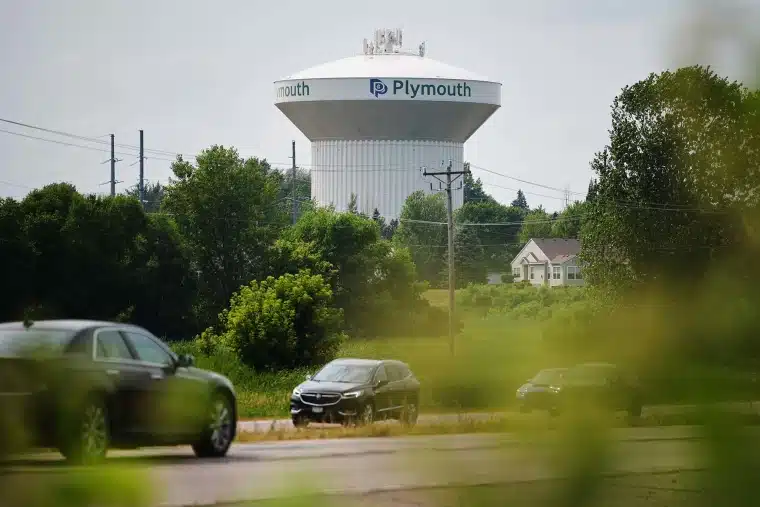 Water tower with Plymouth branding rises above a suburban area, seen behind trees and parked cars on a road in the foreground.